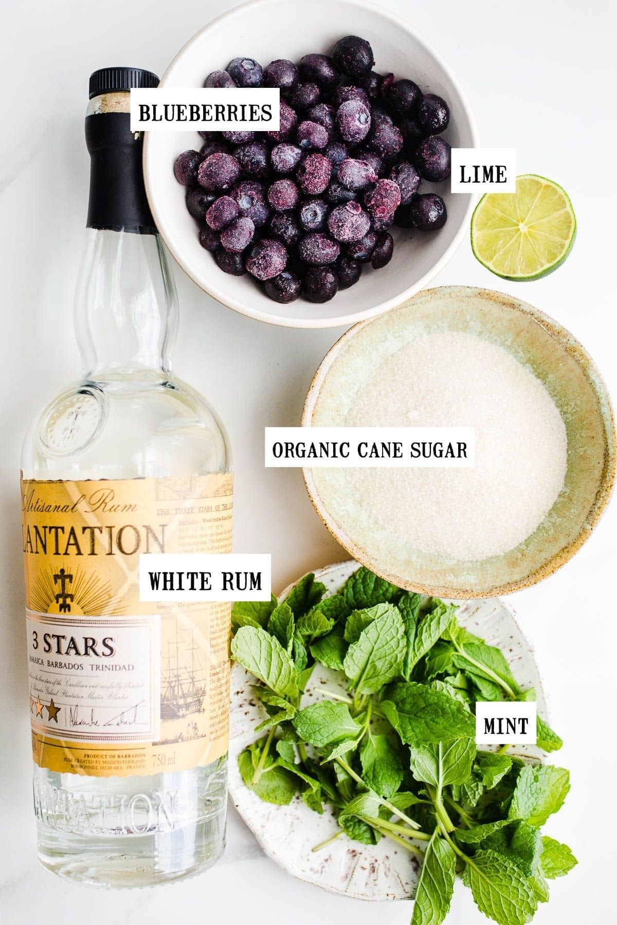 Blueberries, lime, white rum, organic cane sugar, and fresh mint laid out on a marble surface.