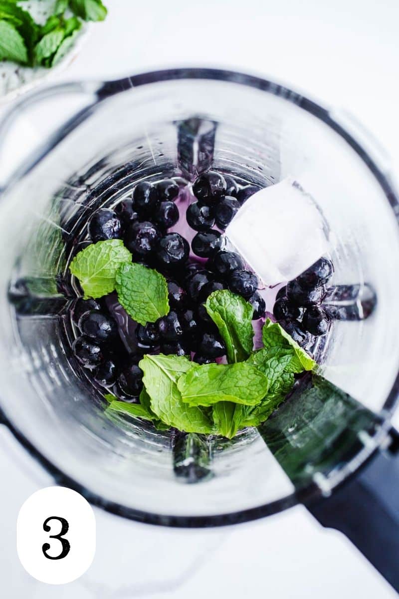 A blender jar containing ice cubes, frozen blueberries, and fresh mint. 