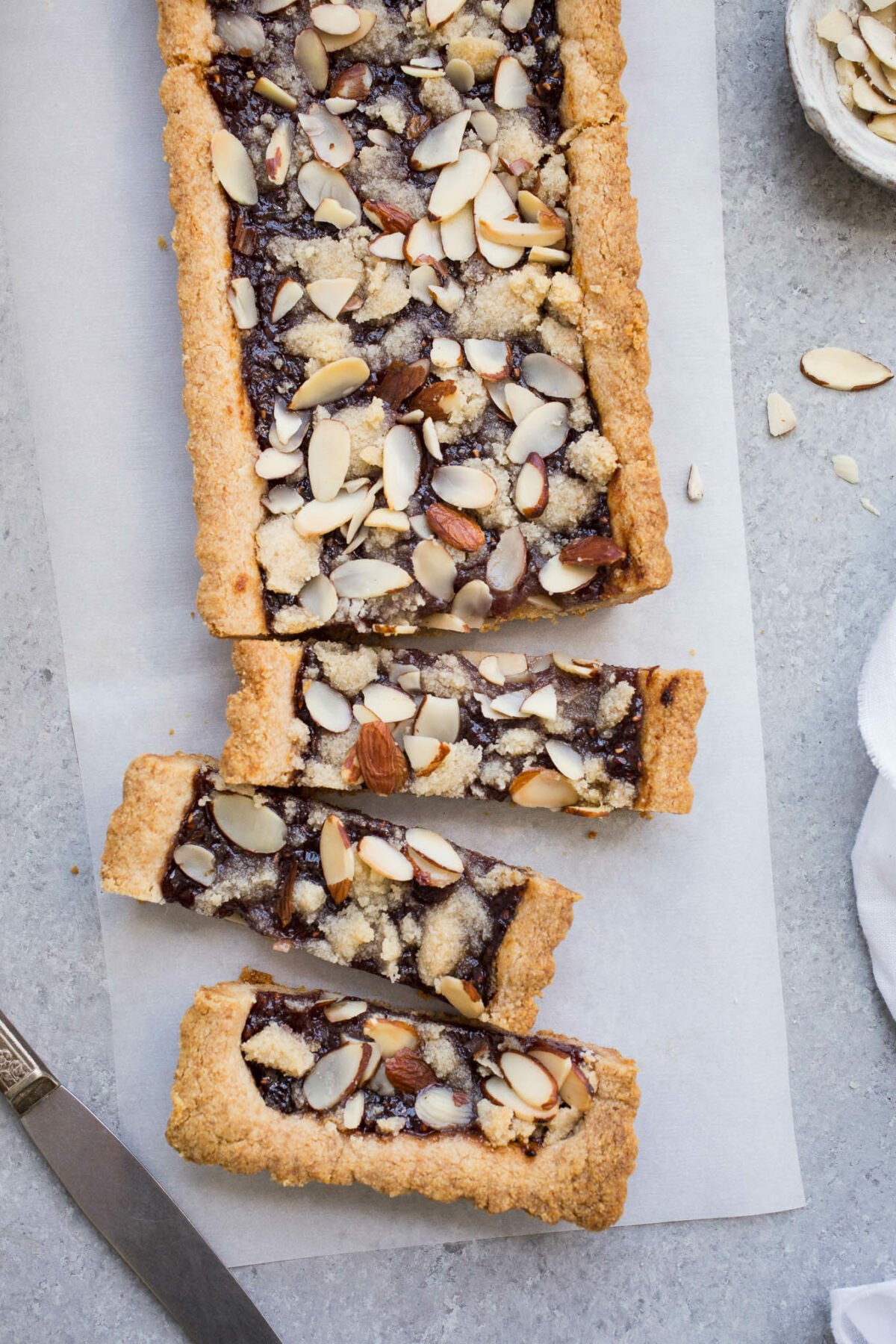 A tart filled with jam and topped with almonds on a piece of parchment paper.