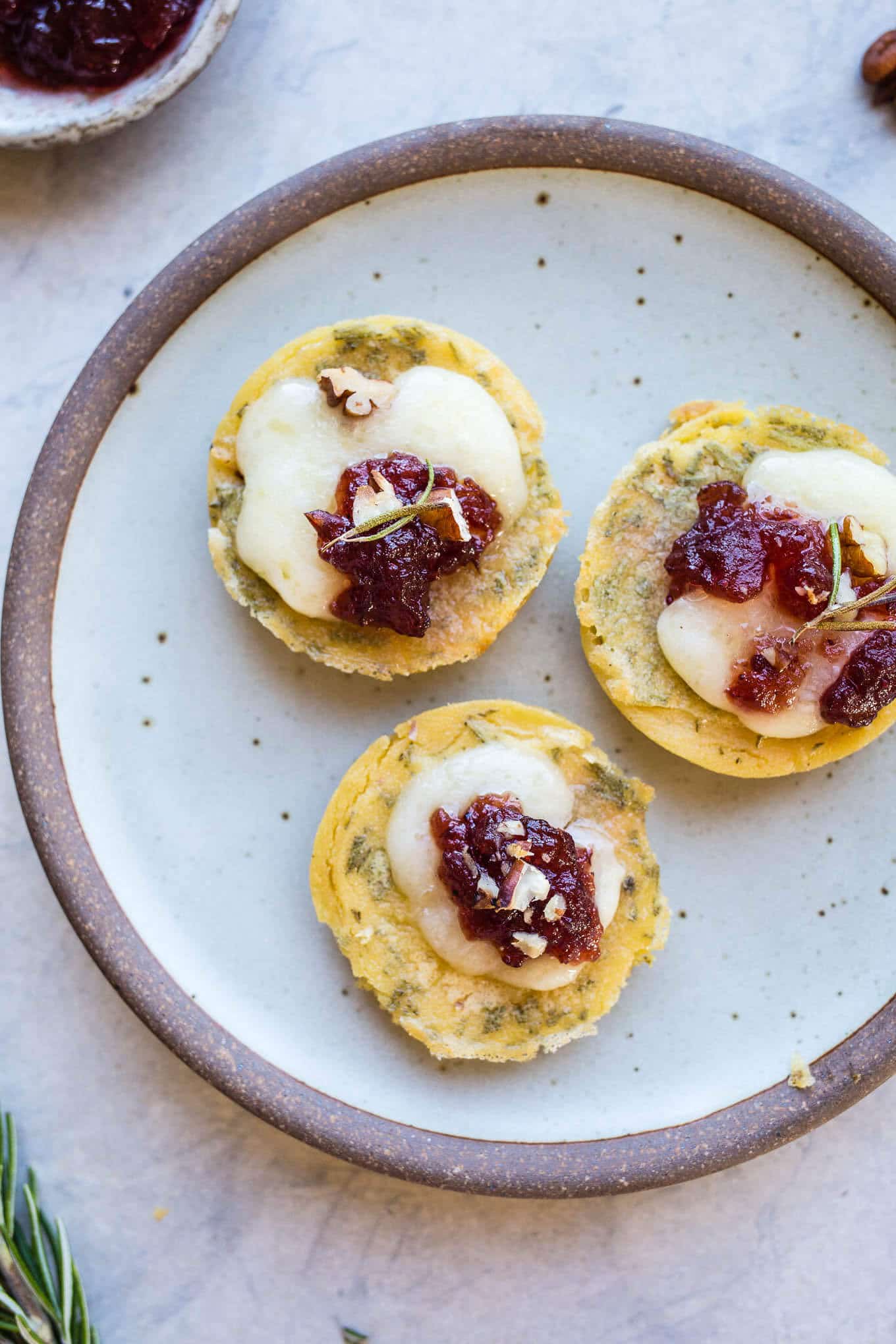 Socca Cranberry Brie Bites are an easy appetizer recipe made from naturally gluten-free chickpea flatbread, brie, cranberry relish, and chopped pecans. Gluten-free.