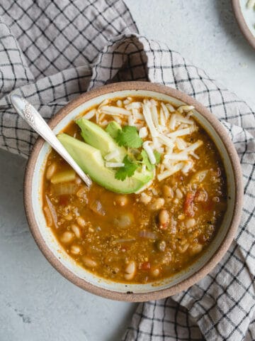 Vegetarian chili in a brown rustic bowl with shredded cheese and sliced avocado on top.