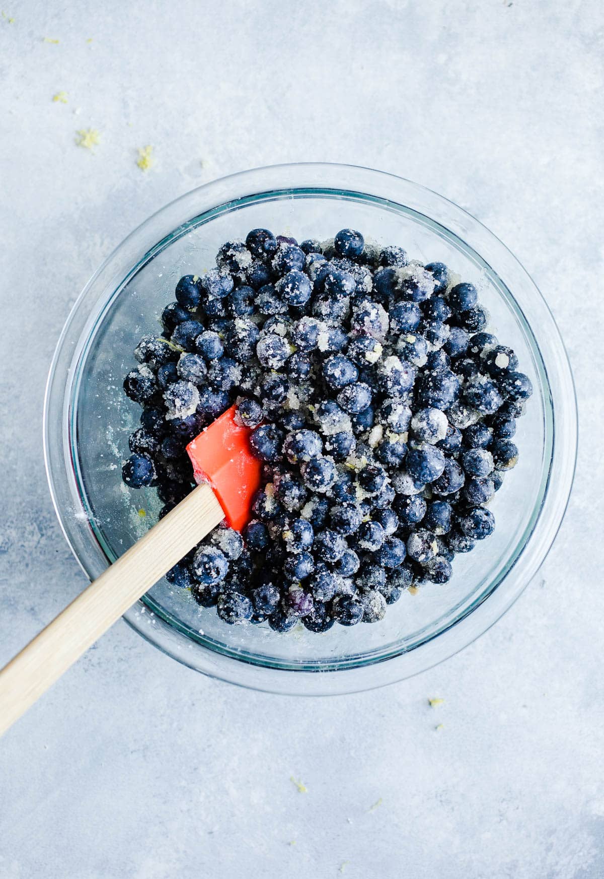 Blueberries, lemon zest, and sugar mixed together in a glass bowl with a red spatula. 