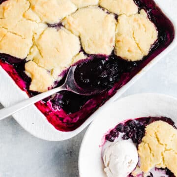 Freshly baked blueberry cobbler in a white baking dish and a scoop of blueberry cobbler in a white bowl with a vanilla scoop of ice cream.