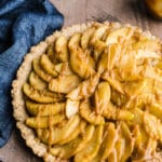 An apple tart drizzled with caramel sit on a wooden surface with a blue cloth.