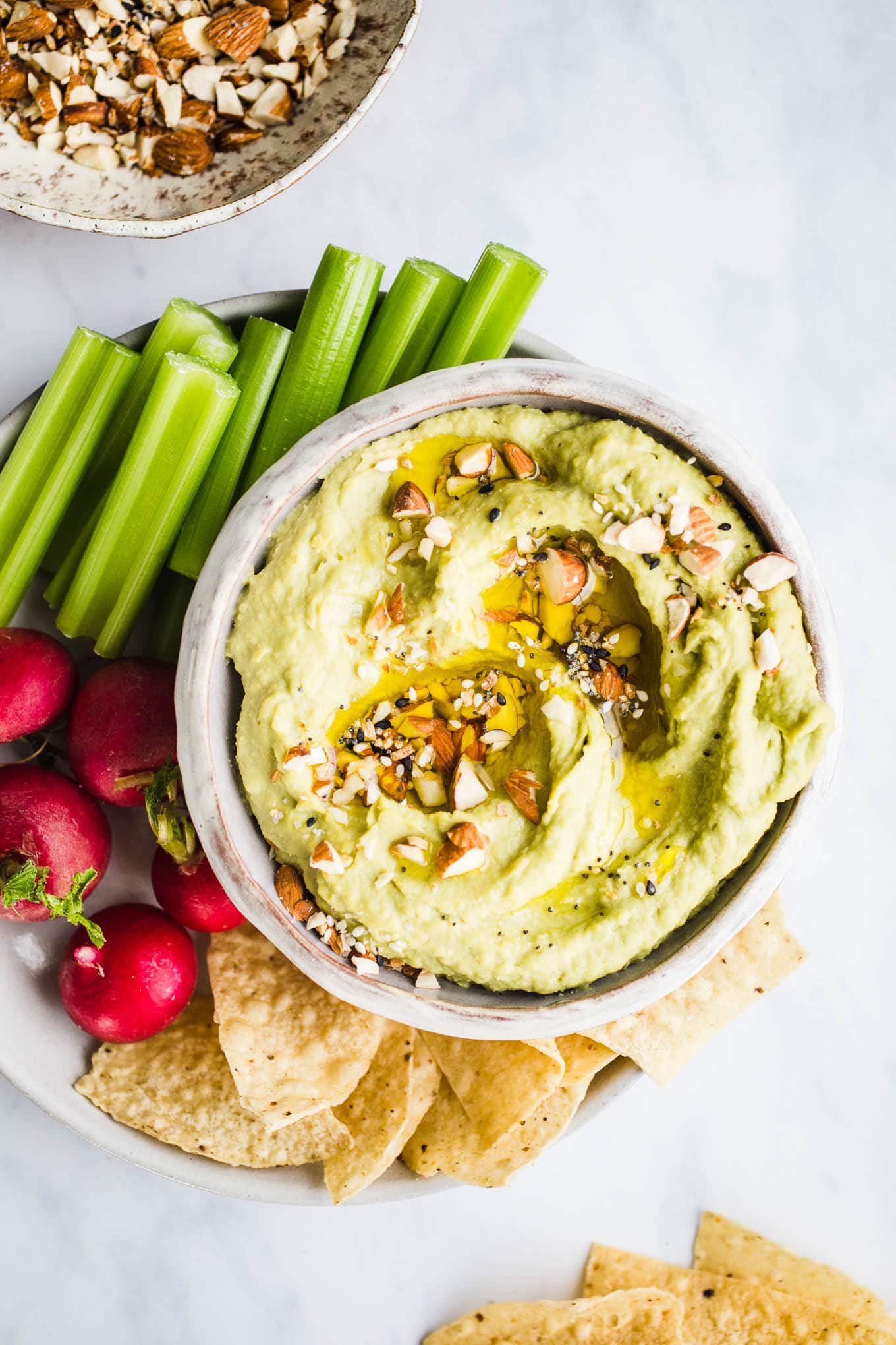 A gluten-free dairy-free snack of green avocado hummus in a white bowl with veggies and tortilla chips.