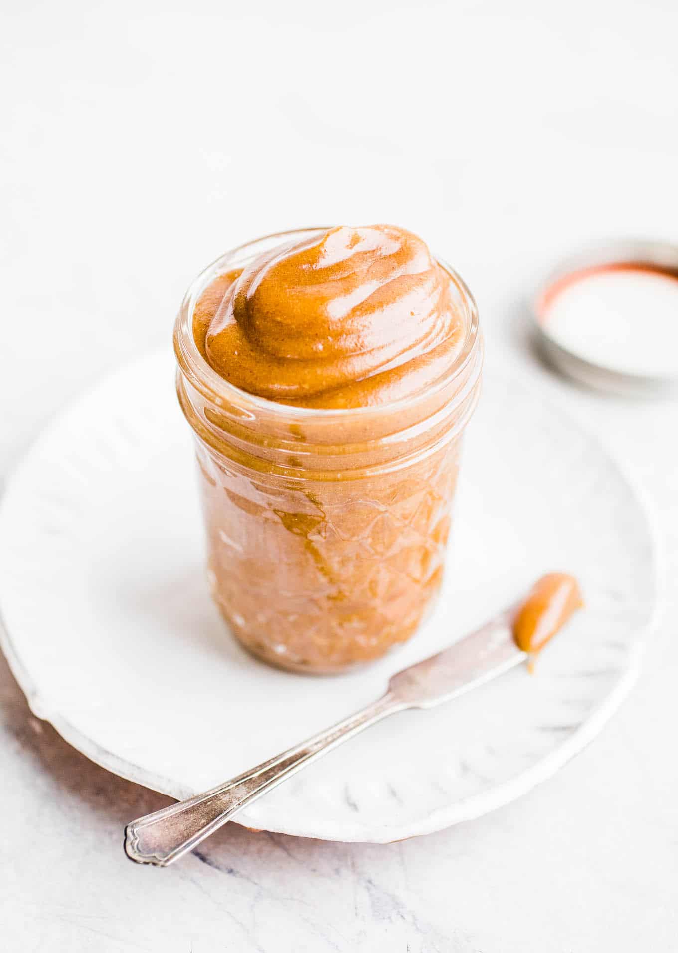 Homemade date caramel in a glass jar sitting on a white plate with a knife that has been dipped in the date caramel.