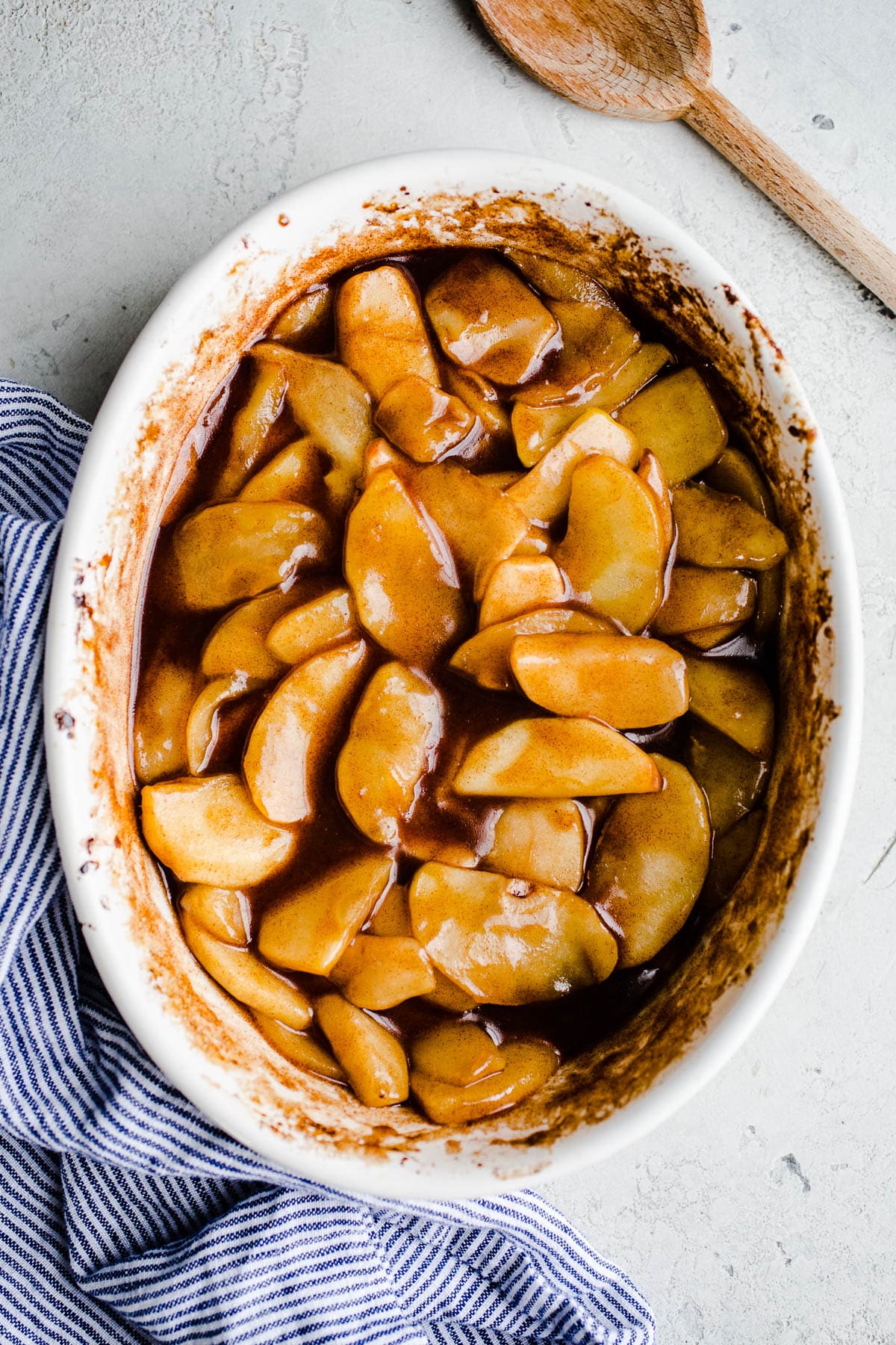 Baked cinnamon apples in a white baking dish next to a striped kitchen towel and wooden spoon.
