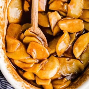 Syrupy cinnamon apples in a white baking dish with a wooden spoon.