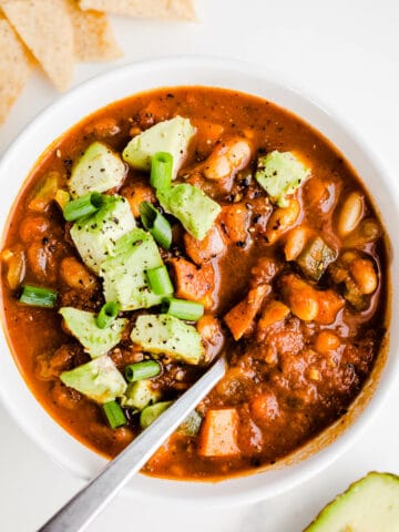 Vegetarian chili with avocado in a white bowl with a silver spoon.