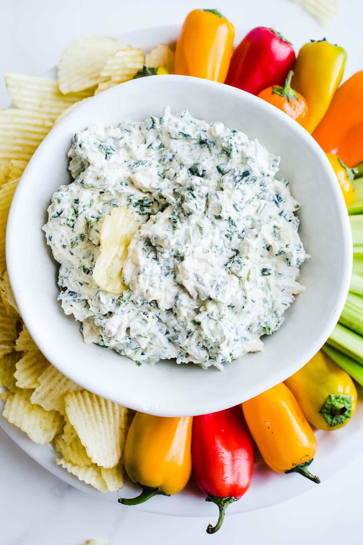 Creamy spinach dip in a white bowl with a potato chip sticking out the middle served with a platter of potato chips, peppers, and celery.