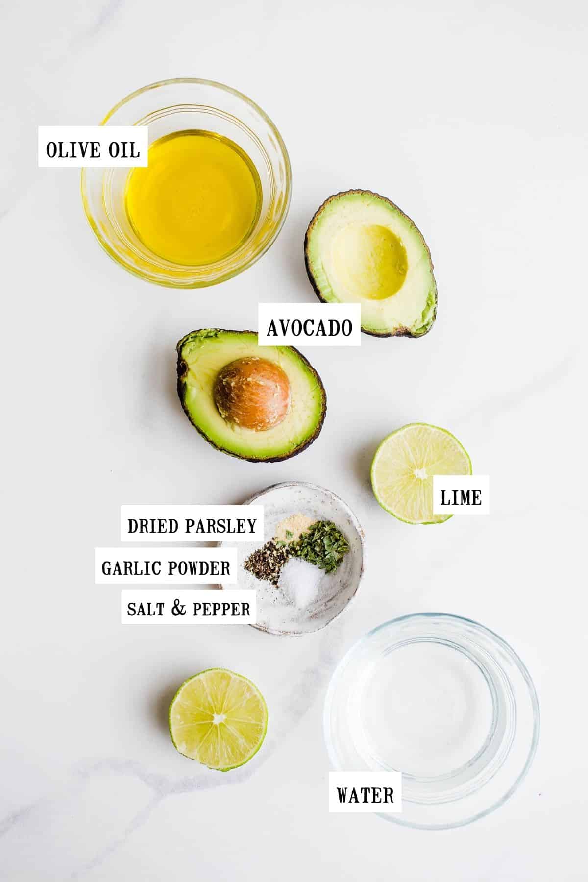 Olive oil, two avocado halves, limes, dried spices, and water laid out on a marble surface.