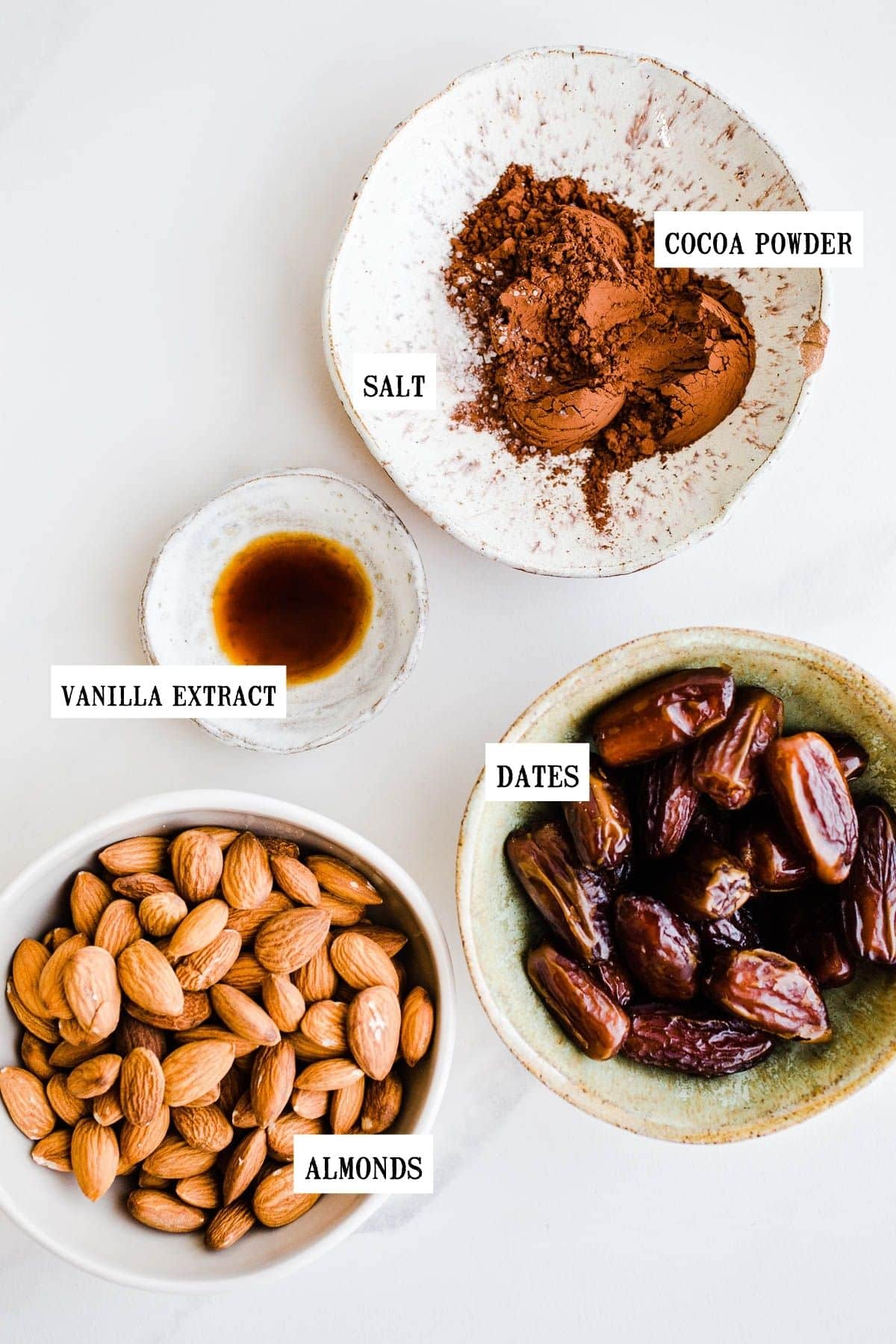 Ingredientes para bolas energéticas en tazones que incluyen cacao en polvo, sal, extracto de vainilla, dátiles y almendras.