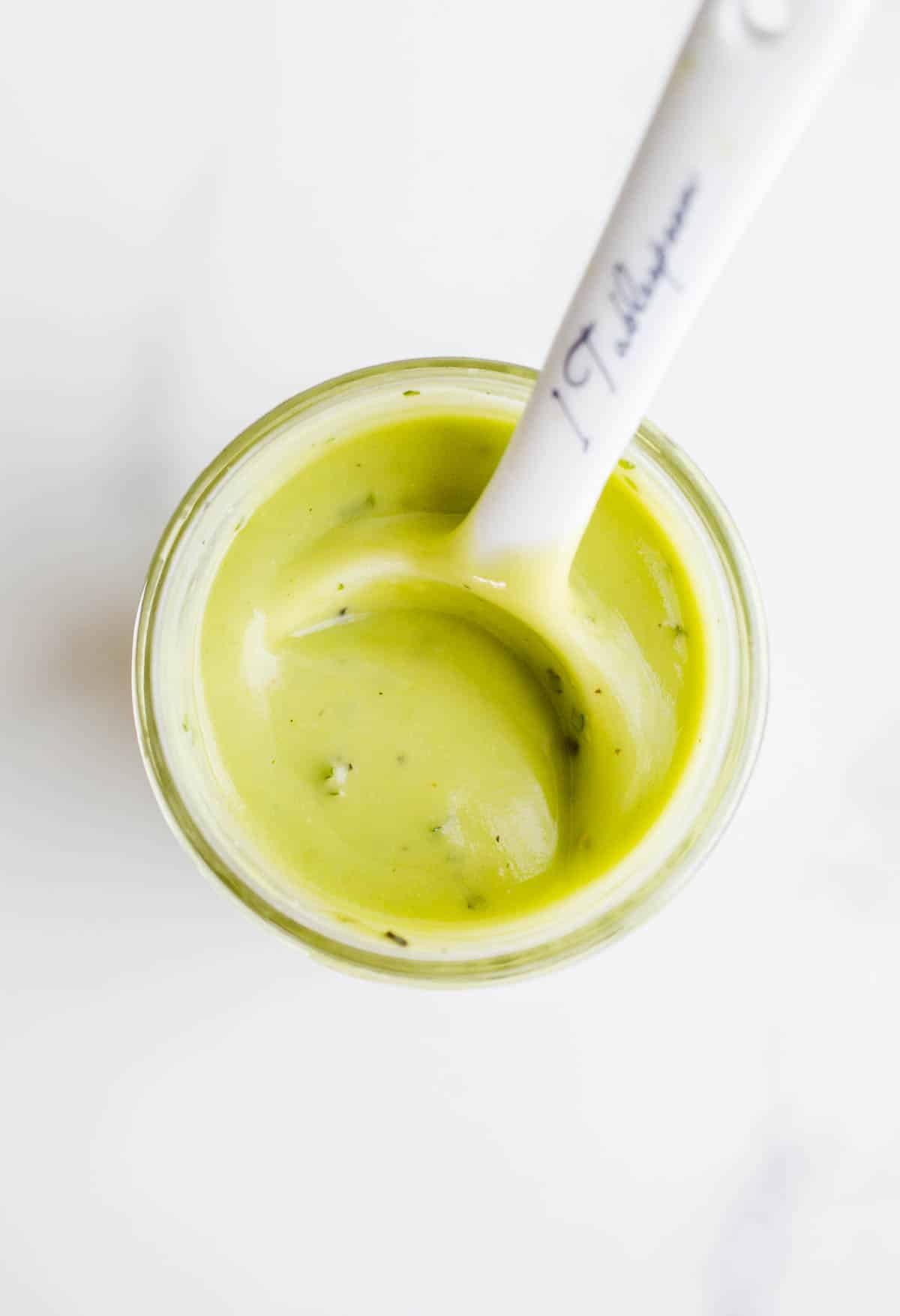 Green and creamy avocado dressing in a glass jar with a white ceramic tablespoon sticking out of it. 
