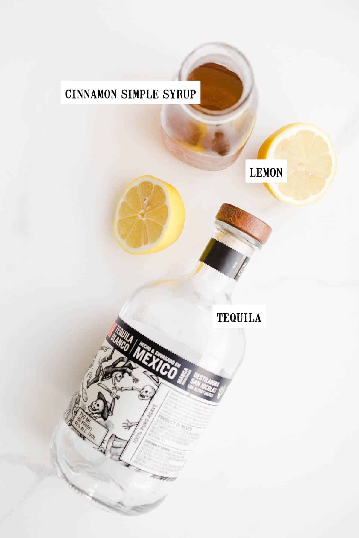 A tequila bottle, lemon, and simple syrup on a white surface. 