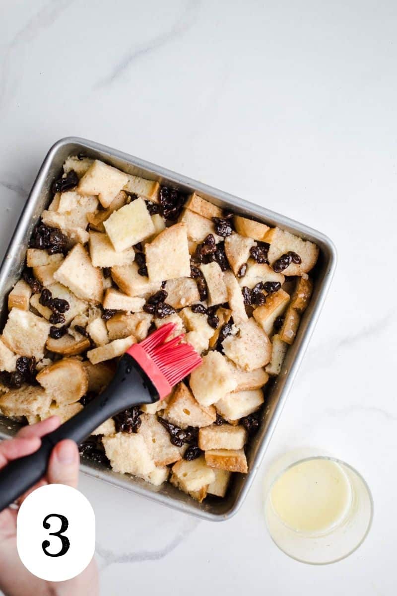 Cubed bread and raisins in a baking pan being basted with vegan butter.