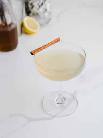 A tequila sour in a coupe glass with a cinnamon stick on a white surface.